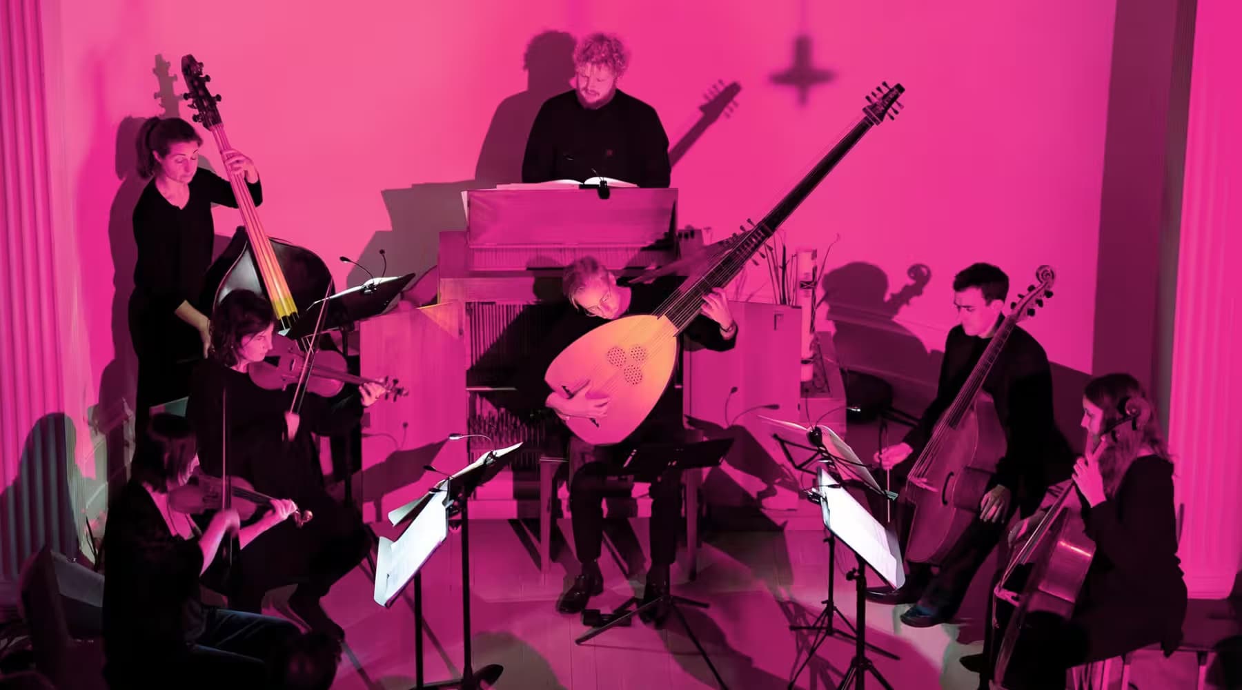 A musical ensemble performs in a room lit with pink light, featuring instruments like lutes, violins, and cellos.